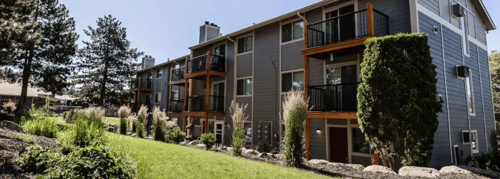 Gray multi-unit apartment building with balconies, lawn, and landscaping.