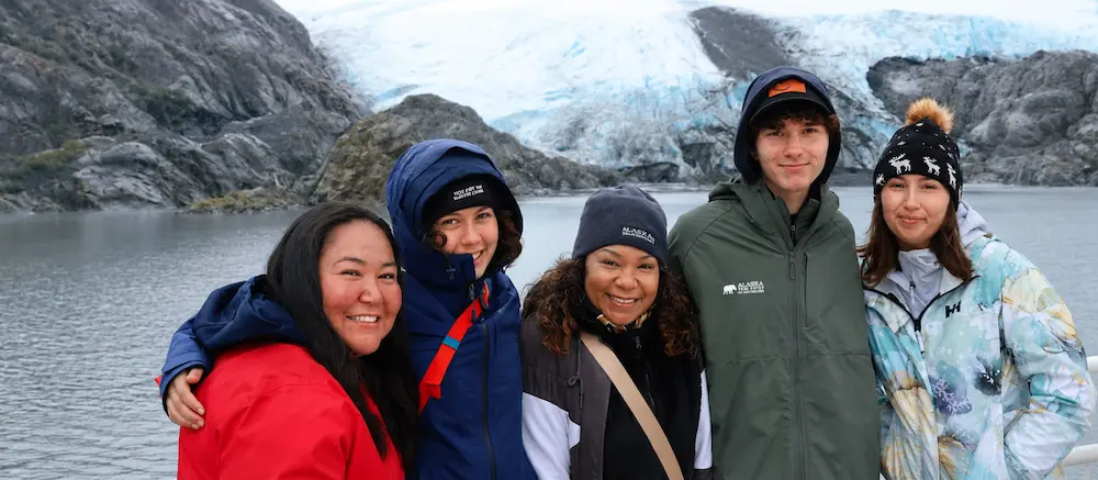 Five people in winter clothing standing beside a glacial lake and ice-covered mountains.