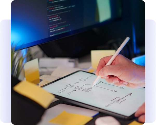 Person using a stylus to draw a workflow diagram on a tablet beside a computer monitor.