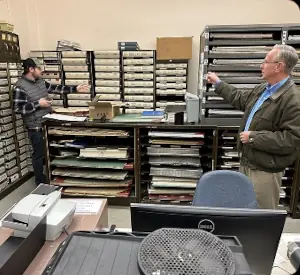 Two employees in an office surrounded by shelves of paper files and materials.